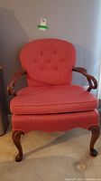 Front view of the pinkish-red upholstered armchair showing cushioned button tufted backrest and carved wooden legs and arms.