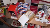 Photo showing boxes of vintage Christmas ornaments, a red collectible box with a Santa figure, and a white snowman plush figurine on a wooden shelf.