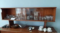 Full view of the wooden shelf displaying various clear glassware including wine glasses, tumblers, and pitchers along with a milk and sugar set on lower shelf