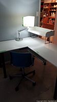 Full view of L-shaped desk with keyboard tray, blue chair, and desk lamp with damaged shade on top of desk.