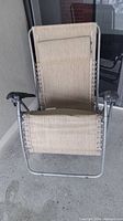 Gravity lounger shown in folded upright position revealing beige fabric seat with elastic cord attachments and metal frame with black armrests.