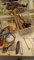 Photo of hand tools such as hammer, utility knives, small hacksaw, pliers and screwdrivers stored in white plastic baskets along with electrical tape and tape measure on a table.