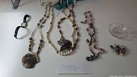 Overview of jewelry including rose quartz necklace, two beaded necklaces with large pendants, wristwatch with black strap, and assorted necklaces