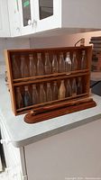 Wooden spice rack holding twenty glass bottles with corks, arranged in two rows on shelves. Bread cutting holder and cutting tool in front.