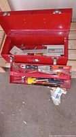 Open red metal toolbox showing internal removable tray with assorted tools inside including various screwdrivers, a hacksaw, pliers, and blades.