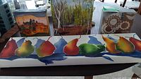 Four pieces of wall art displayed on a table featuring colorful pears, scenic tree paintings, and a decorative wood panel.
