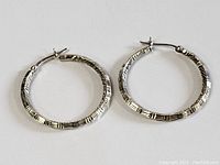 Pair of carved sterling silver hoop earrings shown laying flat on white background. Visible geometric carving detail around hoop.