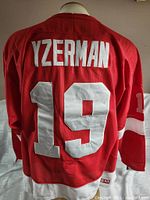Back of bright red hockey jersey showing large white stitched name YZERMAN and number 19, with CCM tag visible on bottom hem.