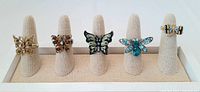 Five rings on a display stand, showing various butterfly and dragonfly designs with rhinestones.