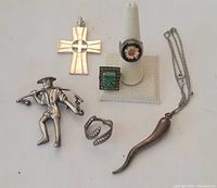 Six pieces of silver jewelry including a cross pendant, necklace with horn pendant, ring with green stone, figurine brooch, and two rings displayed on white surface.
