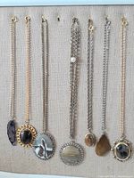 Seven pre-owned natural stone necklaces laid out on beige fabric. Mix of gold and silver chains with varying pendant sizes and shapes.