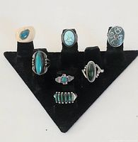 Front view of all 7 rings displayed on a black ring holder stand showing variety of turquoise and other stones in silver settings.