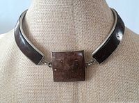 Front view of the necklace on a display bust showing the large square stone centerpiece and cuff style sides with dark stone inlays and silver edges.