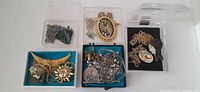 Overhead view of assorted costume jewelry pieces in three small boxes and one plastic container, showing mixed silver-tone pendants, chains, charms and green faceted beads