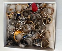 Overhead view of a box filled with assorted rings of various metals and designs including silver, gold-tone, and colored stones.