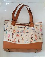 Front and part of side view of Disney tote bag, showing cream background with colorful Disney characters and brown handles and trim.