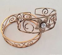 Pair of vintage brass open cuff bracelets with decorative spiral motifs, one featuring a flower with rhinestone centerpiece.