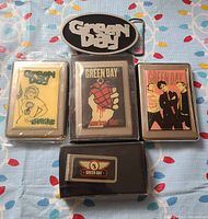 Green Day belt buckle, 3 cigarette cases, and 1 money clip laid out on patterned surface