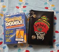 Photo of Seeing Double playing cards next to black wallet with skull and 'SMOKE' embroidery and cannabis leaf