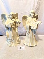 Front view of two cream ceramic angel candlestick holders, the violin-playing angel on the left and harp-holding angel on the right, showing overall size and design.