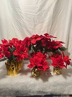 Six artificial poinsettia plants with red petals and green leaves, each in gold foil wrapped bases, arranged on a white surface.