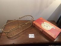 Wire bread basket with gold-tone metal wire in a chicken-wire pattern on wooden surface alongside the cheese knife set box.