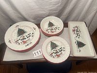 Four Christmas-themed ceramic dishes including dinner plates, salad plates, bowls, and rectangular serving tray with holiday tree and village design.