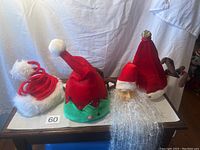 Four Christmas-themed hats and one Santa decoration: red twirly spiral hat, green elf hat with bells, red reindeer hat, and Santa head hanging decoration with beard.