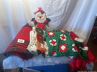 Photo showing the entire lot stacked on a surface: red and black plaid tablecloth, white patterned tablecloth, green crocheted Afghan shawl, brown round table runner, two sequin stockings (green and red), and NOEL Santa Claus wall hanging.
