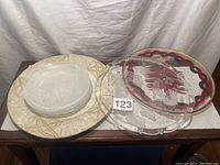 Four etched glass plates, one etched glass serving tray, a Christmas tree serving platter with gold trim, and a cream and gold decorative plate displayed on a wooden surface.