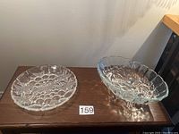 Photo showing two clear crystal items on a wooden table: a round serving plate with floral etching and an oval dish with scalloped edges and floral pattern.
