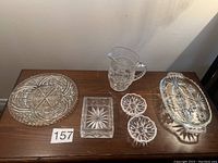 Full lot view of three candy dishes, two crystal coasters, and one water jug on wooden table.