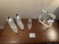 Two ceramic decorative high heel shoes, small glass decorative shoe, and clear glass vase on a wooden surface with lot number 168.