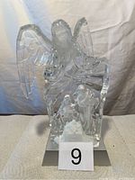 Front view of a clear hard plastic angel holding a musical instrument with a manger scene beneath on a silver base.