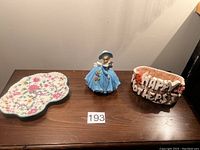 Three items on a wooden surface: floral candy dish plate, lady figurine, and Happy Mothers Day plastic basket
