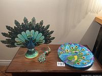 Photo showing three peacock items: decorative plate, ornament, and candle holder arranged on a table.
