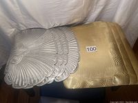 Four gold colored square place mats with embossed wave design stacked with four silver colored fan-shaped place mats stacked alongside.