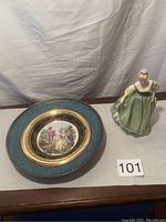 Photo showing the Royal Doulton Fair Lady figurine next to the decorative plate on a flat surface with plain background.