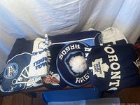 Toronto Argonauts and Toronto Maple Leafs apparel and accessories folded on table including t-shirts, scarf, towel, and hats