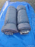 Two rolled grey sleeping bags with plaid lining set on blue tarp outdoors, showing thickness and outer design.