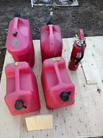 Four red plastic gas cans and one red fire extinguisher arranged on a wooden surface outdoors