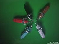Top-down view of five crystal pendants arranged in a star pattern on a dark surface. The metal capped pointed crystals show various colors: pale blue, white, red, black, and reddish-brown.