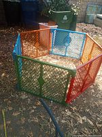 Collapsible playpen set up outdoors on ground covered with leaves and dirt, showing multi-colored connected plastic panels forming a hexagon shape