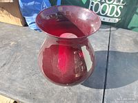 Large ruby red glass hurricane lamp with wide flared top and rounded body shown on outdoor surface, with visible candle inside.