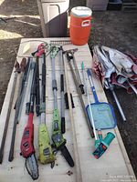 Overview photo of various used garden tools including pole saws, hedge trimmer, pruners, net, long wooden and metal poles, large orange water cooler jug, and folded fabric pieces on a wooden pallet.