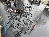Photo showing three bicycle-shaped metal plant stands on wet pavement. The largest is in the rear left with large basket and decorative wheels.