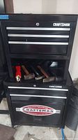 Full view of black metal Craftsman toolbox with 5 drawers and wheels. Several hammers and a pruning shear laying on open bottom shelf.