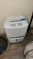 Frigidaire white dehumidifier against a beige wall and on a wood floor, with power cord visible on the floor.