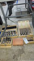Five drill bit sets arranged on floor with metal stand behind