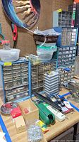 Photo showing various metal organizer cabinets stacked with small hardware drawers, plastic bins, and assorted hardware items on a workbench beneath a pegboard wall.